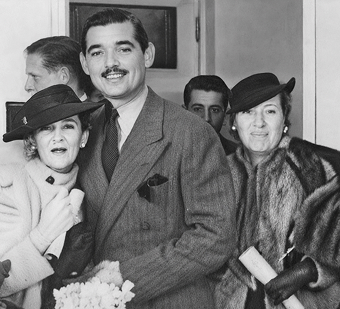 Black-and-white photo of a smiling man in a suit between two women wearing hats and fur coats at an indoor gathering.