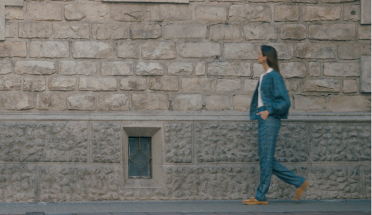Woman in a teal jacket walks past an old stone wall with a small barred window.