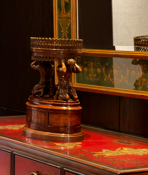 Ornate antique wooden pedestal with carved figures stands on a red marble tabletop in a richly decorated room.