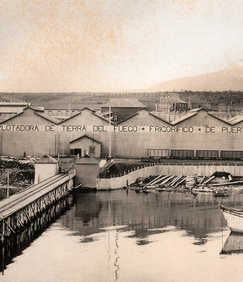 Sepia photograph of a waterfront meatpacking plant in Tierra del Fuego, with docks, moored boats, and a long industrial building.