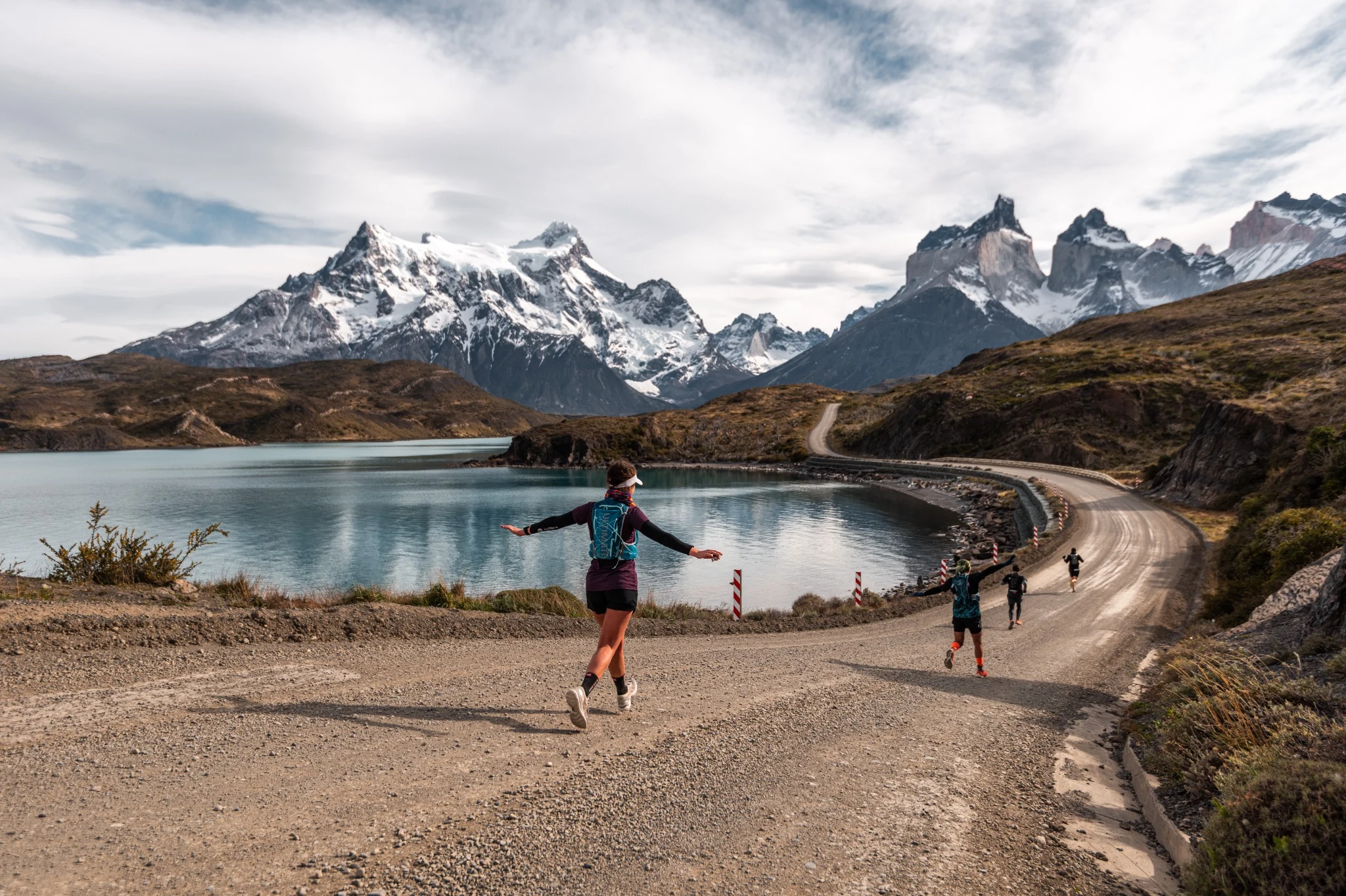 Trail runners follow a winding lakeside road beneath snow-capped Patagonian peaks.