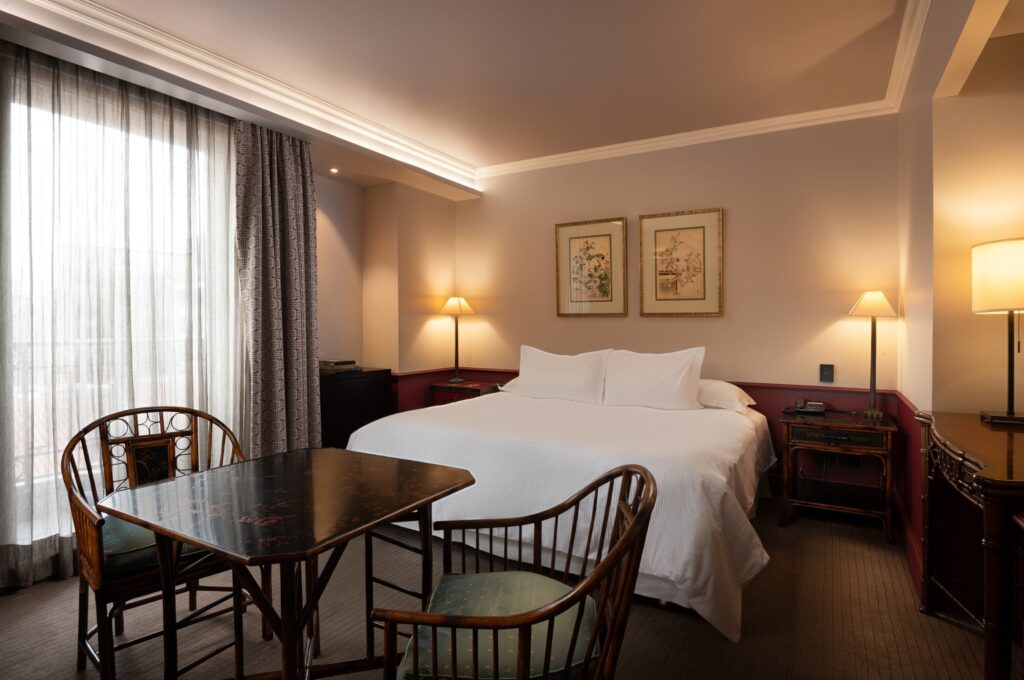 Elegant hotel room with a king bed, antique dining table and chairs, framed art, warm lamps, and sheer-curtained windows.