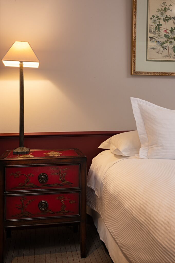 Cozy bedroom with a glowing table lamp on an ornate red bedside chest beside a neatly made bed and framed floral art.