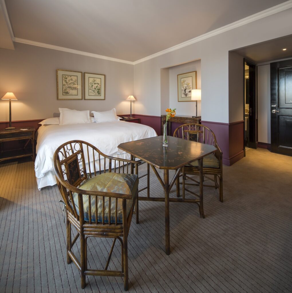 Elegant hotel bedroom with a white bed, antique wooden dining table and chairs, framed botanical art, and warm lamplight.