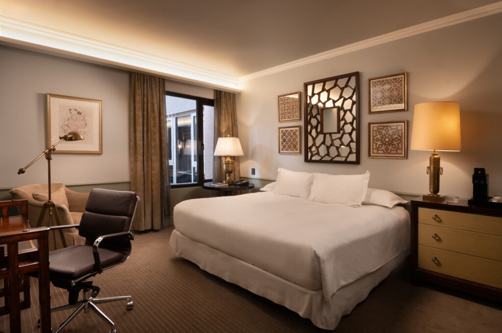 Elegant hotel bedroom with a king bed, warm lighting, framed geometric art, a desk, and a seating area.