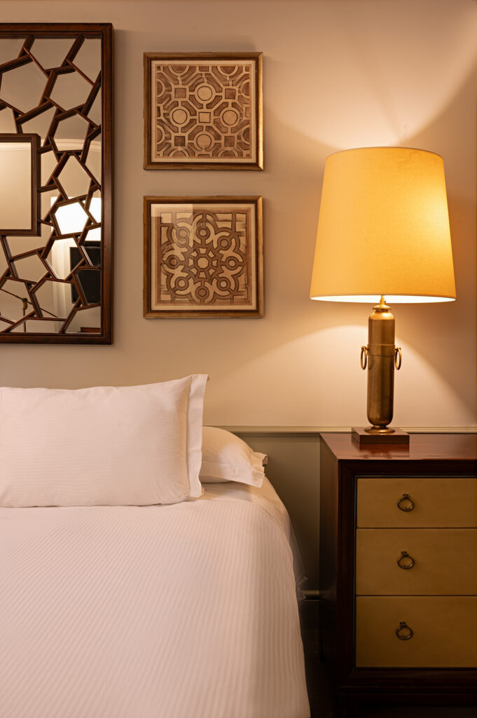 Cozy bedroom with white bed, glowing brass table lamp, geometric framed artwork, and dark wood nightstand beside a decorative mirror.