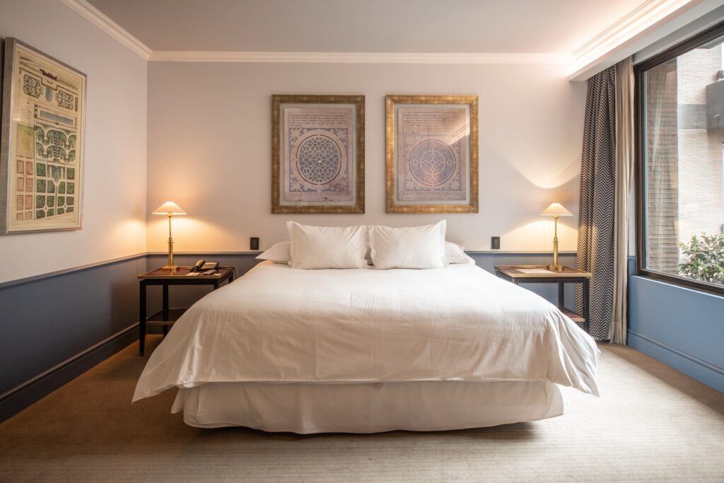 Elegant hotel bedroom with a king bed, framed artwork, bedside lamps, and a large window with patterned curtains.