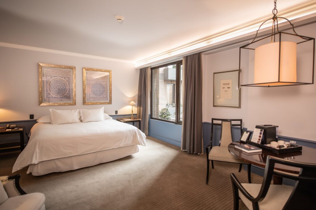 Elegant hotel bedroom with a king bed, framed artwork, large window, patterned curtains, lounge chairs, and a glowing pendant light.