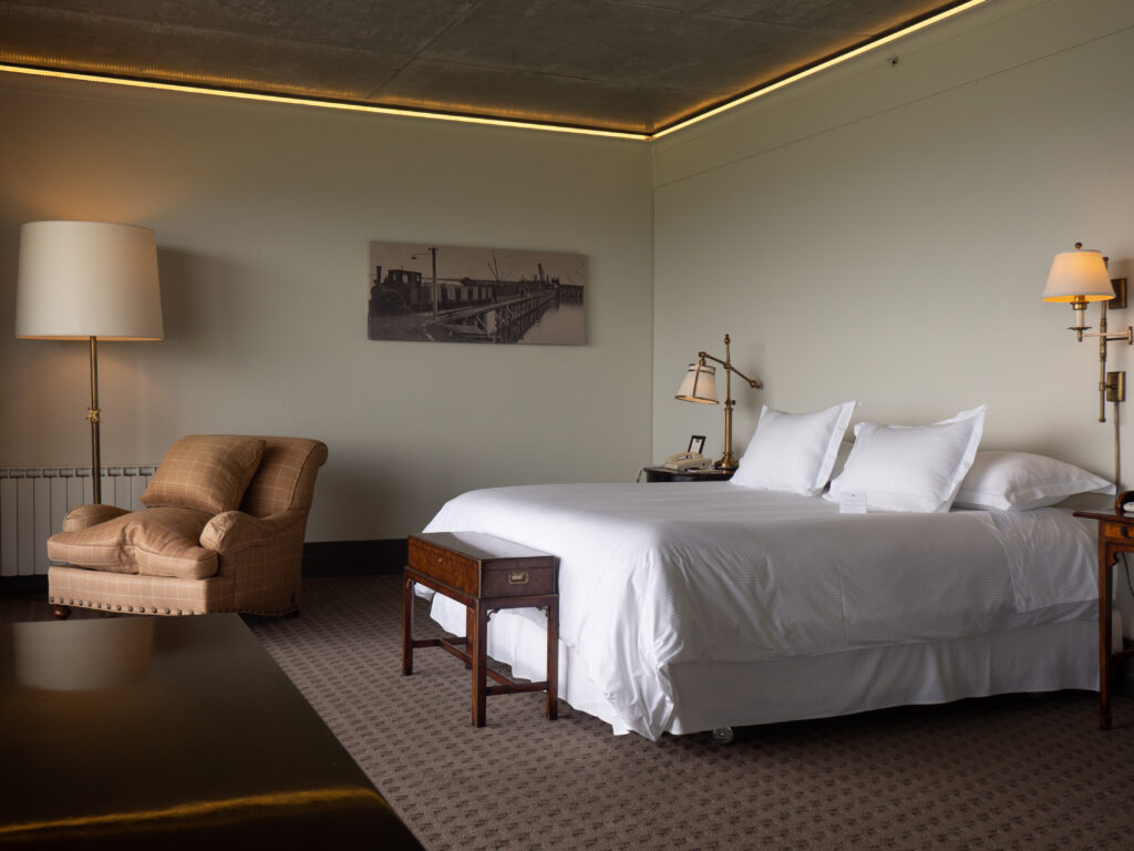 Elegant hotel bedroom with a white bed, tan armchair, antique wood furniture, warm lamps, and historic railway artwork.