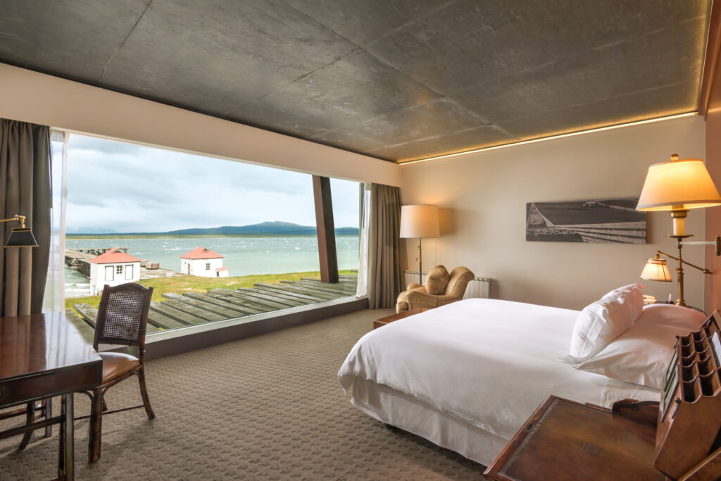 Modern hotel bedroom with a panoramic window overlooking a lake, dock, and distant mountains.