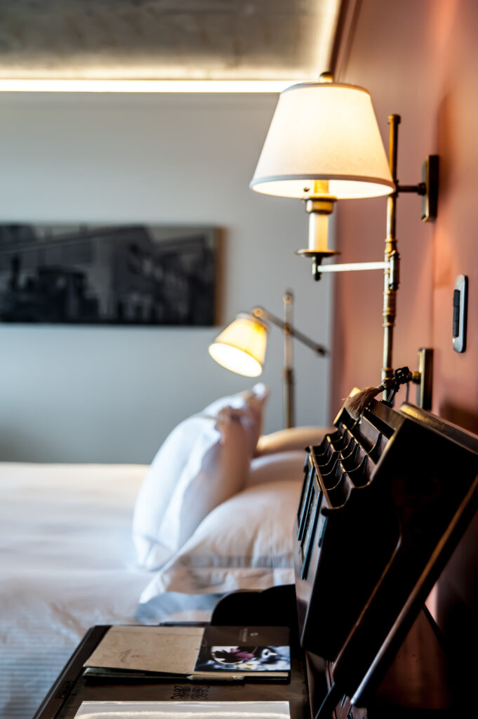 Warm bedside wall lamp illuminates a neatly made bed, dark headboard, and bedside table in a cozy bedroom.