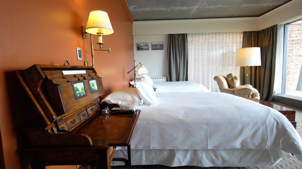 Elegant hotel room with two white beds, antique wooden desk, warm lamps, and a lounge chair beside large curtained windows.