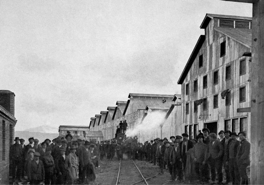 Black-and-white historical photo of workers lining railway tracks beside textile mill buildings as a steam locomotive arrives.