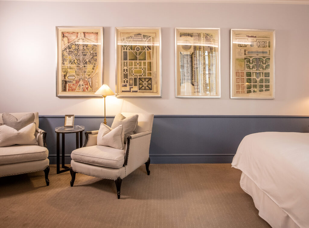 Elegant lounge with cream armchairs, a lit floor lamp, and four framed antique garden plans on the wall.