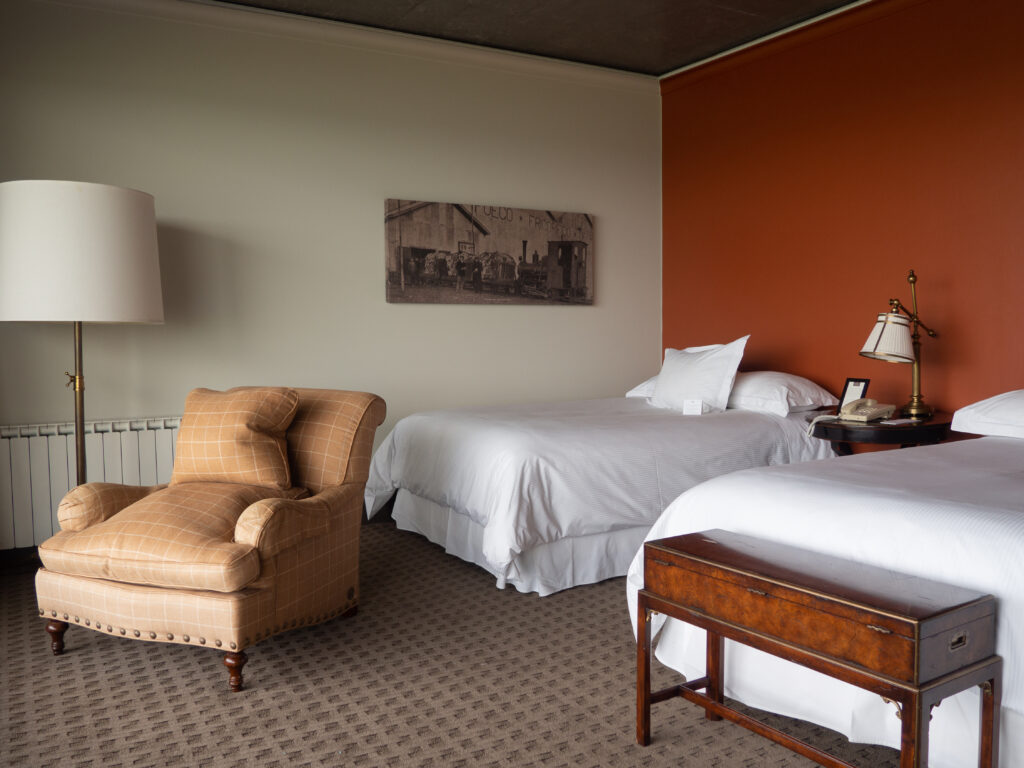 Hotel room with two white beds, a tan armchair, bedside lamps, antique wooden luggage bench, and rust-red accent wall.