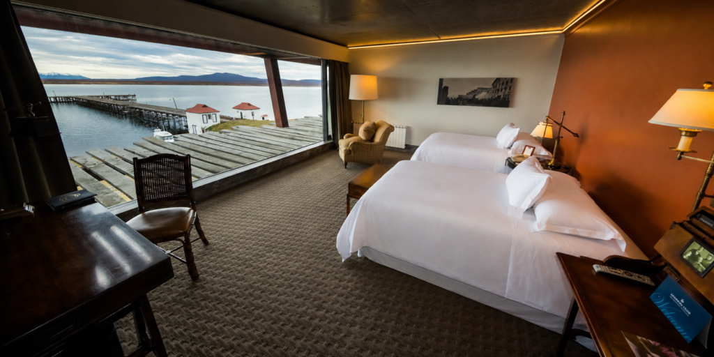 Hotel room with two beds and floor-to-ceiling windows overlooking a lake, dock, and distant mountains