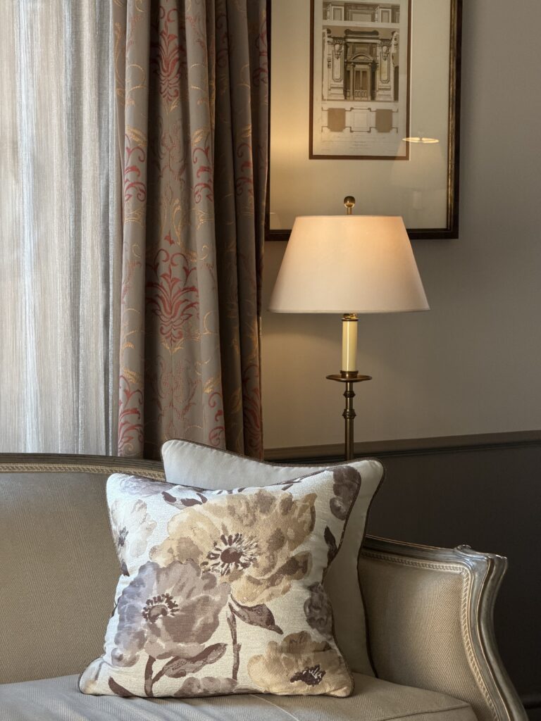Traditional living room with floral pillows on a beige sofa, patterned curtains, a brass floor lamp, and framed architectural art.