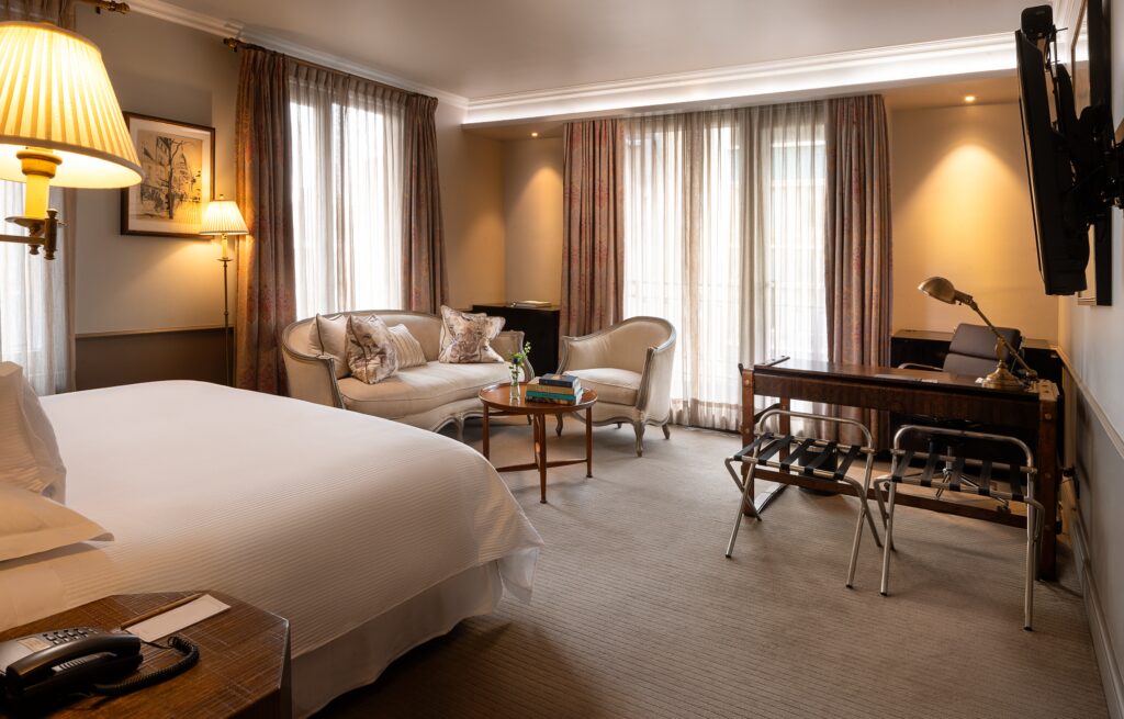 Elegant hotel suite with king bed, sofa seating, writing desk, large curtained windows, and warm lighting.