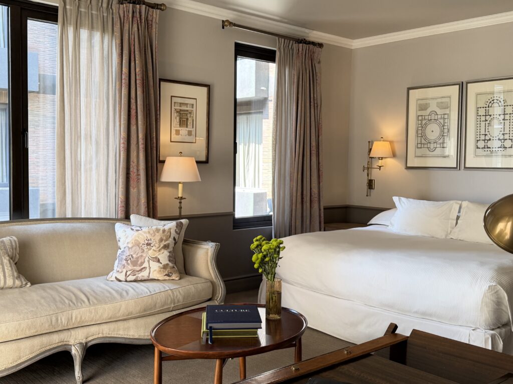 Elegant hotel bedroom with white bed, cream sofa, framed architectural prints, tall curtained windows, and a flower vase on a coffee table.