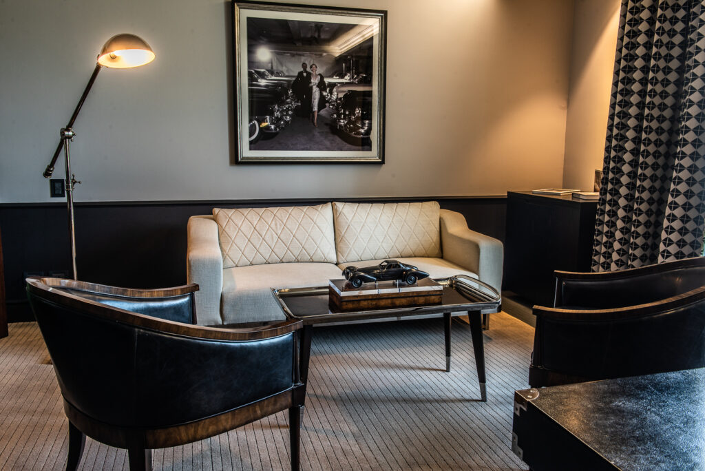 Elegant hotel lounge with cream sofa, dark leather chairs, vintage car model, floor lamp, and framed black-and-white photograph.