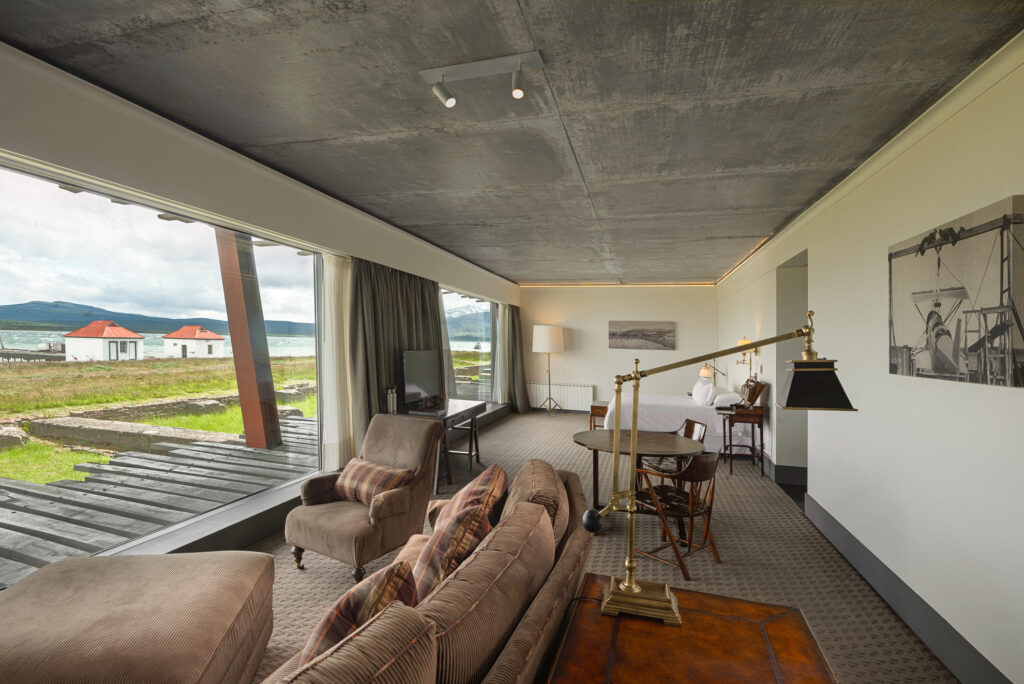 Modern hotel suite with a bed, lounge seating, and floor-to-ceiling windows overlooking a grassy lakeside landscape.