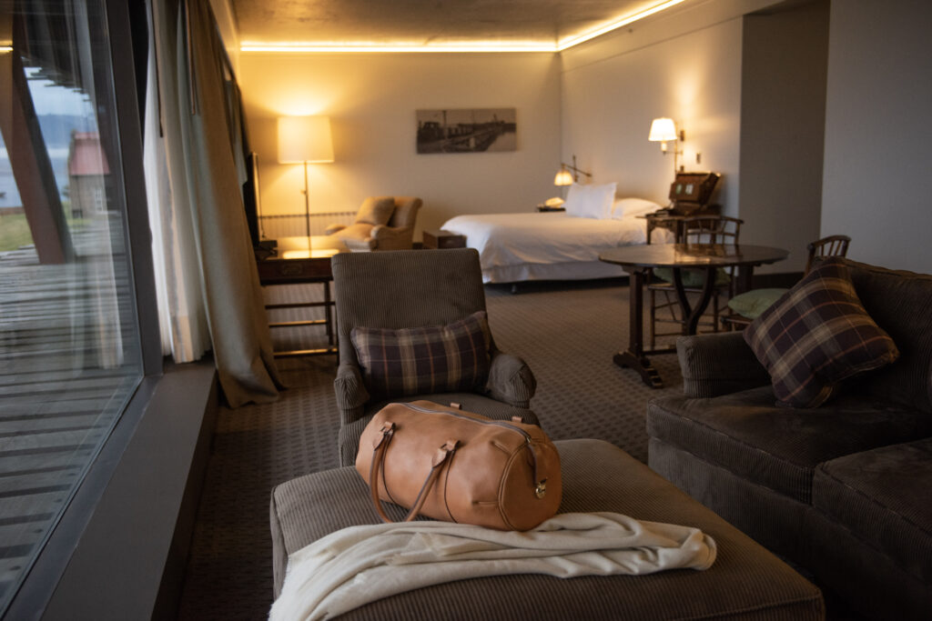 Warmly lit hotel suite with a king bed, lounge chairs, a window-side balcony, and a leather travel bag in the foreground.