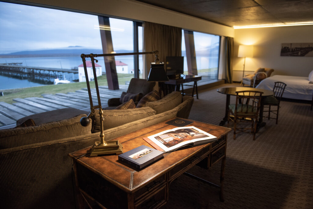 Cozy hotel suite with vintage furnishings, a waterfront view through large windows, and a bed beside a seating area.