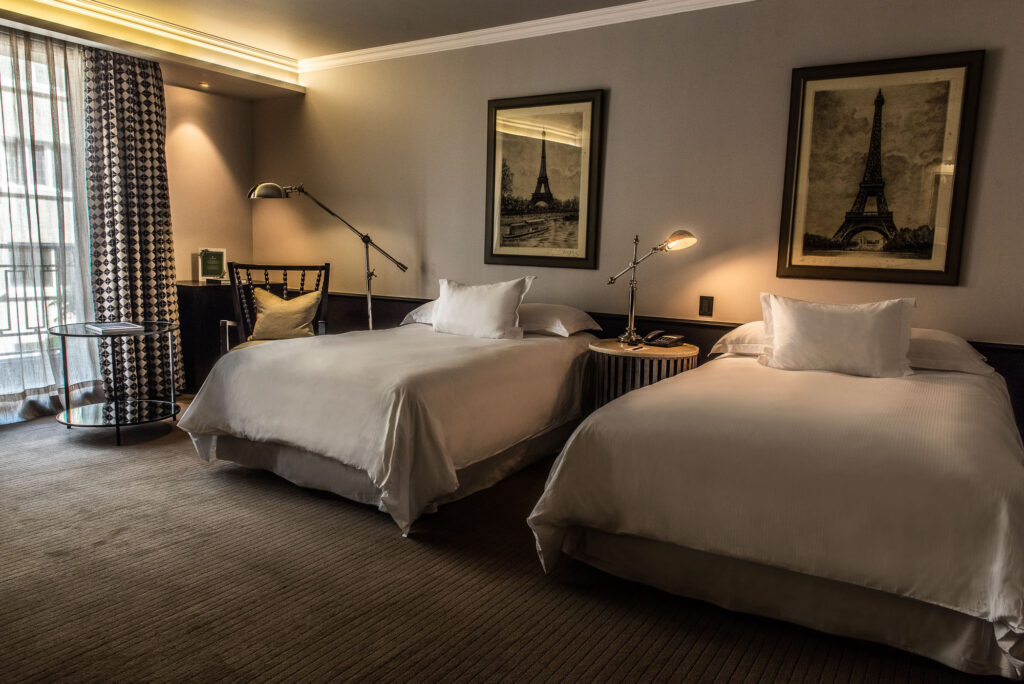 Elegant hotel room with two white beds, warm bedside lamps, patterned curtains, and framed Eiffel Tower prints.