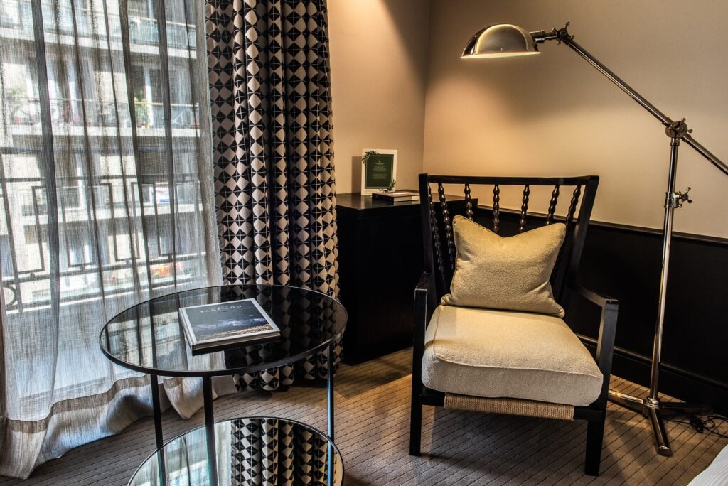 Elegant hotel room sitting area with a beige armchair, brass floor lamp, glass table, and patterned curtains by a window.