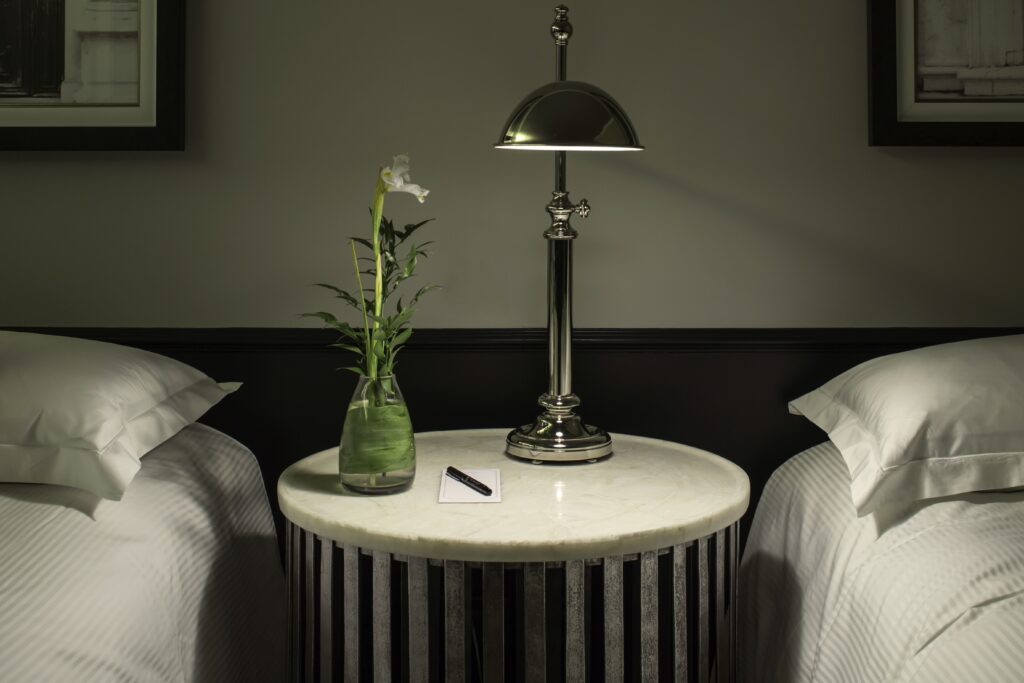 Hotel room bedside table with a polished lamp, white flower in a glass vase, notepad, and pen between two beds.
