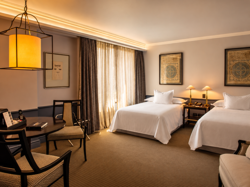 Elegant hotel room with two white beds, warm bedside lamps, a round dining table, and sunlit curtains.
