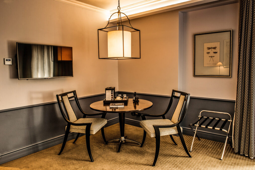 Elegant hotel room sitting area with round table, two upholstered chairs, wall-mounted TV, pendant light, framed artwork, and patterned curtains.
