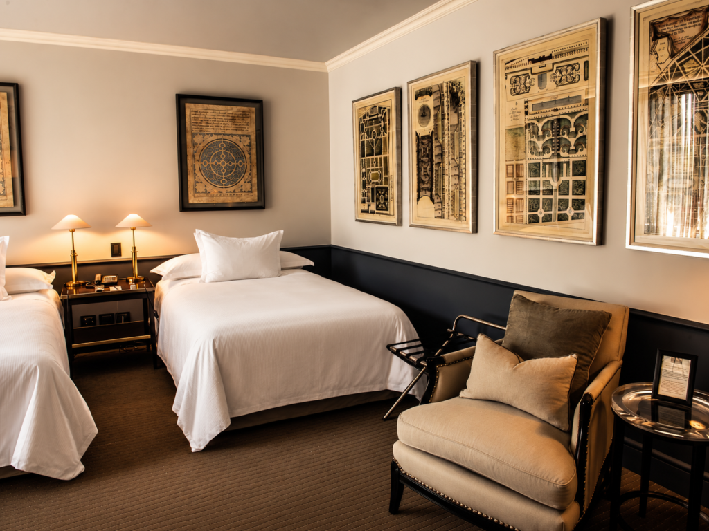Elegant hotel room with two white beds, framed artwork, warm bedside lamps, and a cushioned armchair.
