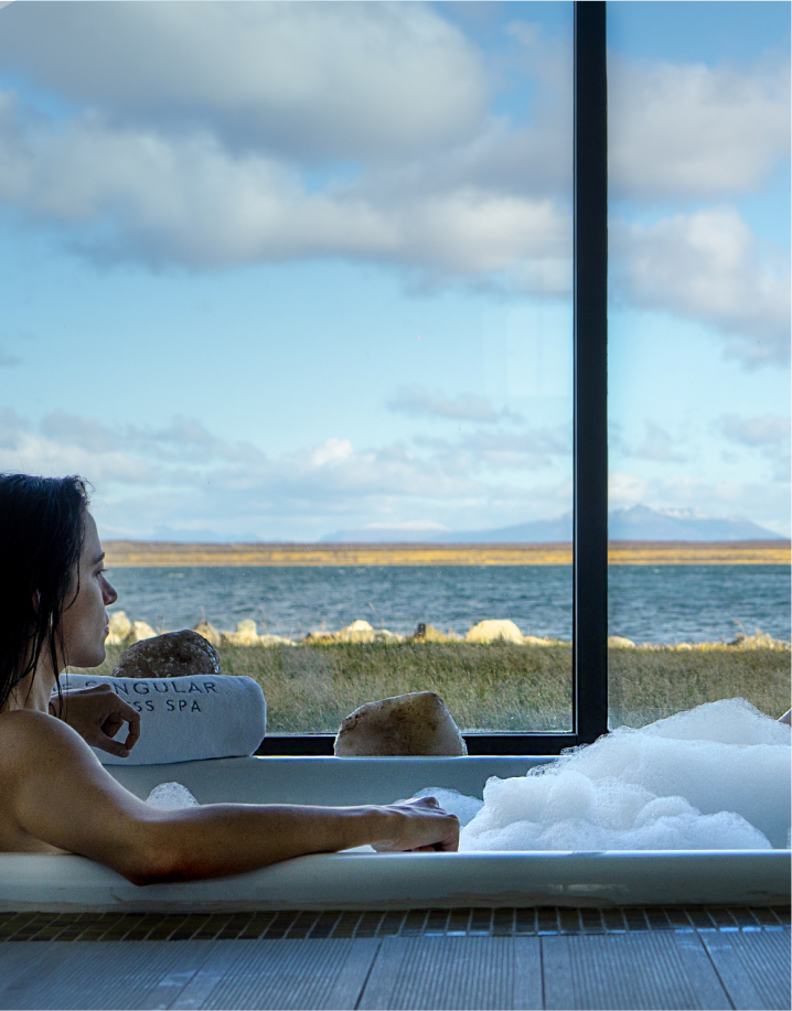 wellness-3 Woman relaxing in a bubble bath beside a large window overlooking a lake and grassy shoreline.