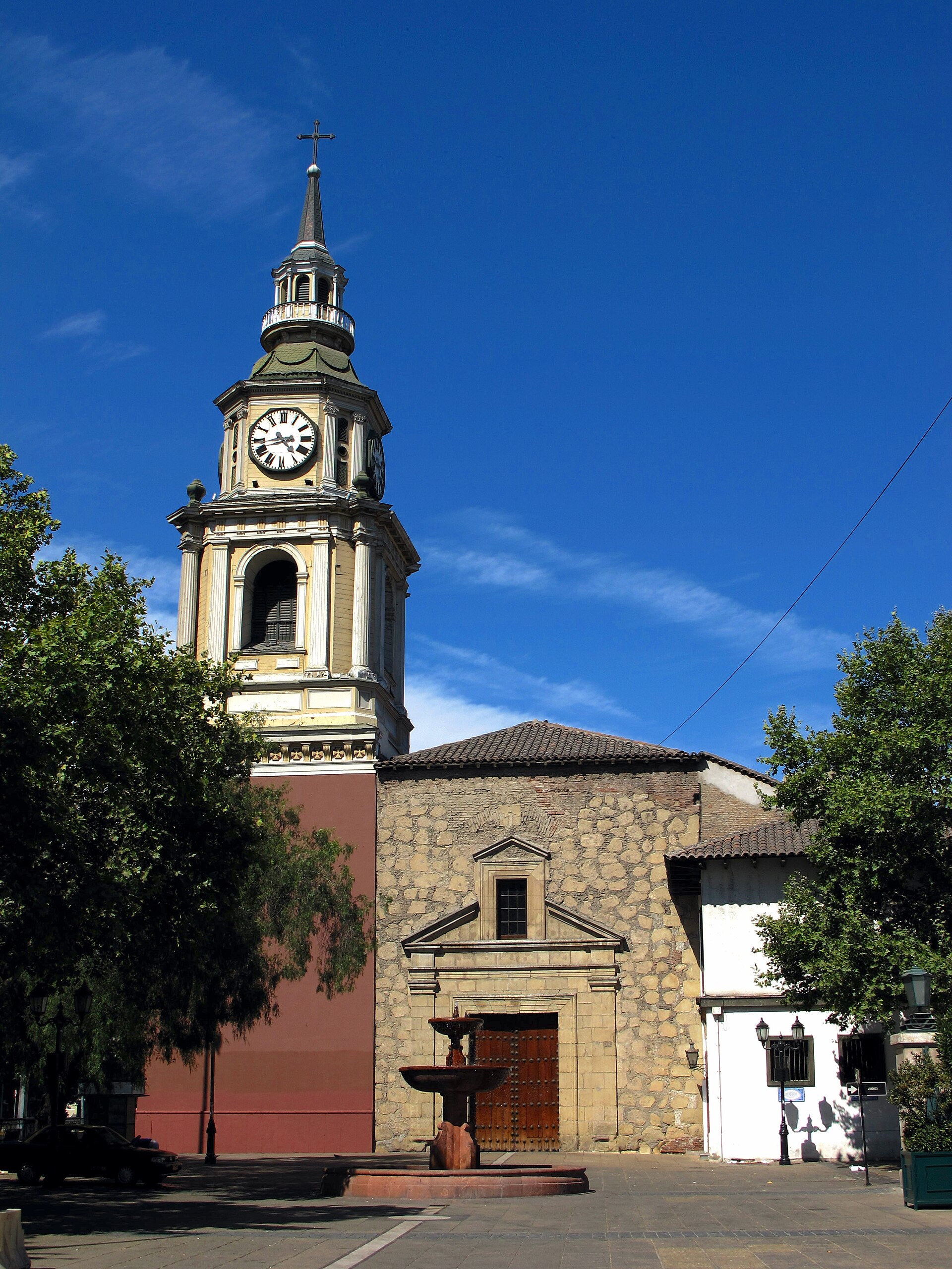 Historic stone church with a clock tower and cross rises above a plaza fountain beneath a clear blue sky.