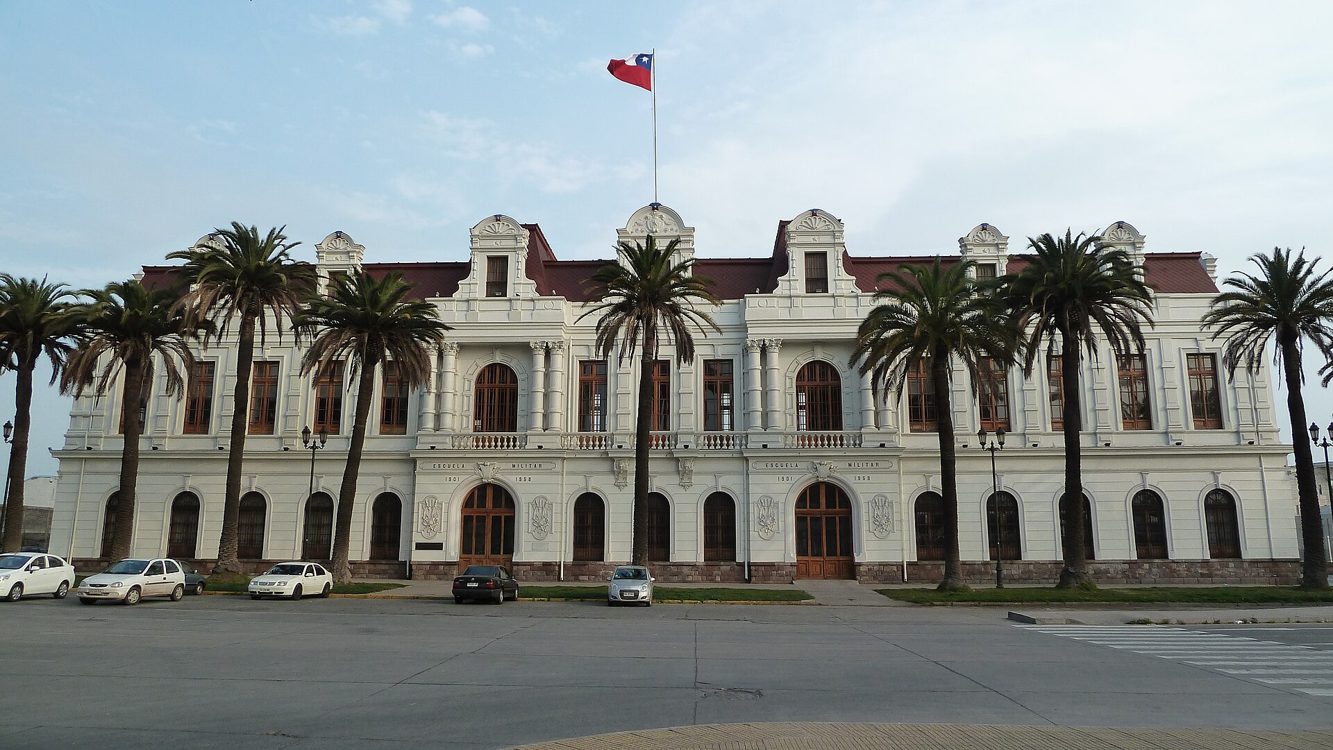 Historic white Escuela Militar building with Chilean flag, palm trees, and parked cars in front in Santiago, Chile.