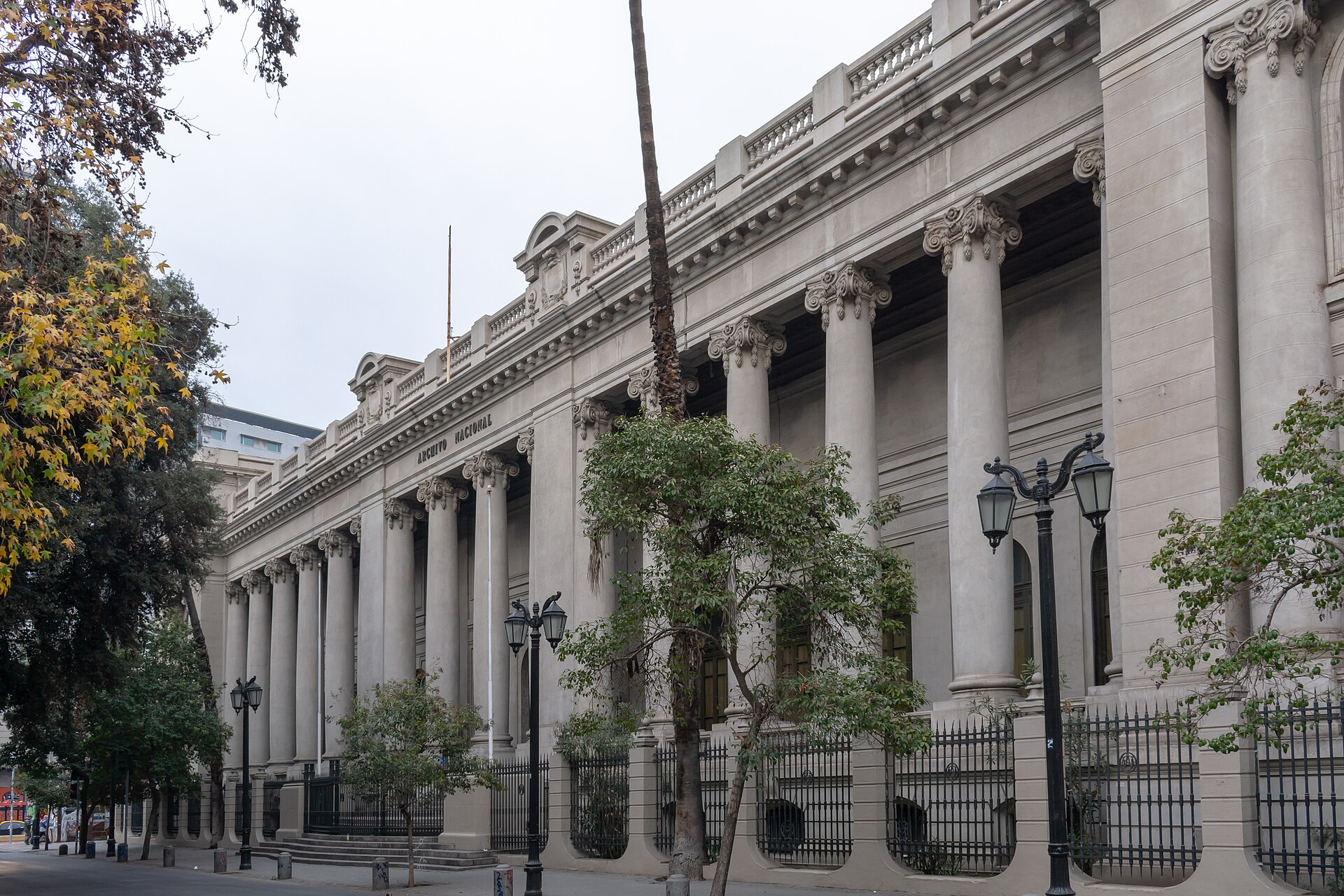 Neoclassical National Archives building with ornate columns, iron fencing, trees, and streetlamps along the sidewalk.