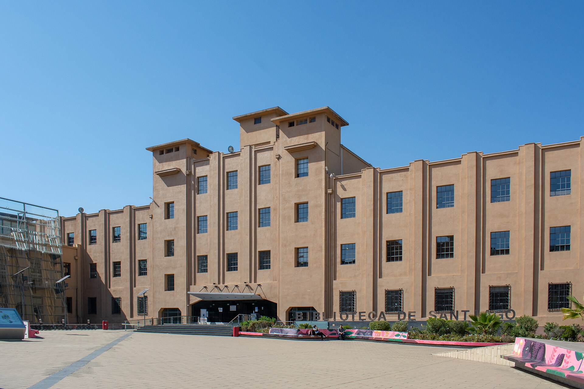 Santiago Library, a large tan modernist building with rows of windows, stands beside a sunny paved plaza.