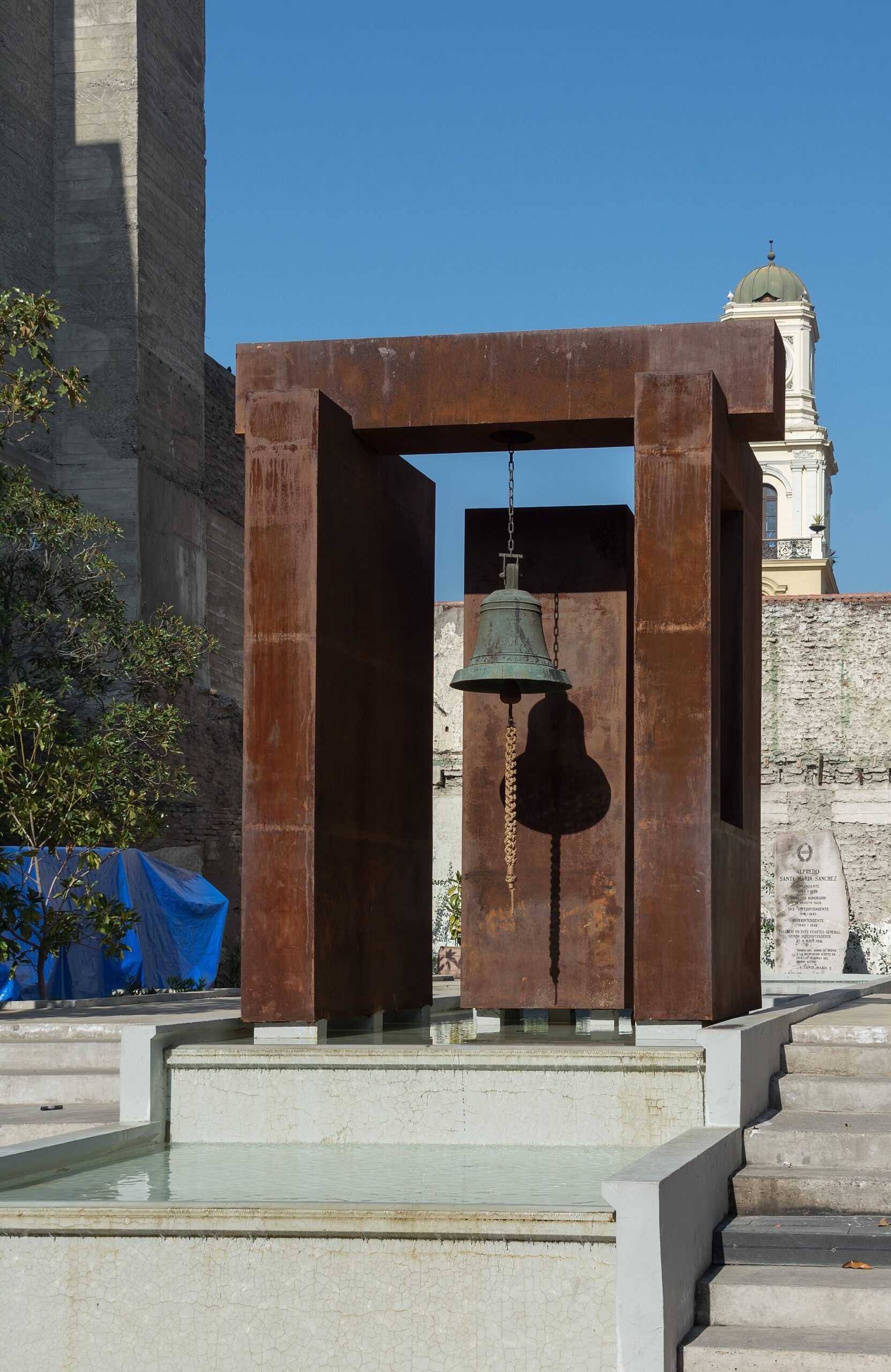 Large bronze bell suspended in a rusted steel monument above a reflecting pool, with a historic domed tower behind it.