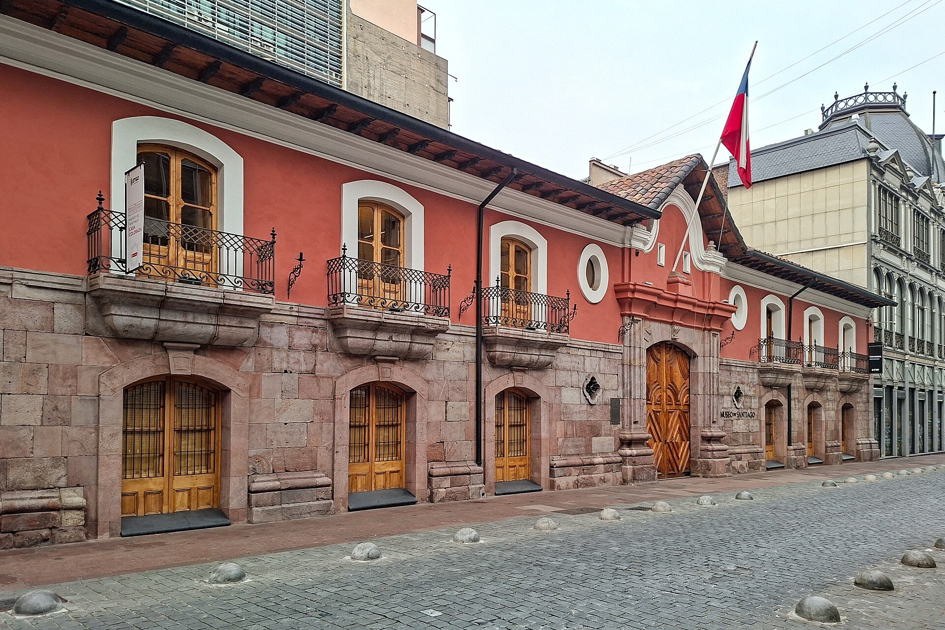 Historic red Casa Colorada museum with stone lower facade, ornate balconies, wooden doors, and a Chilean flag on a cobblestone street.