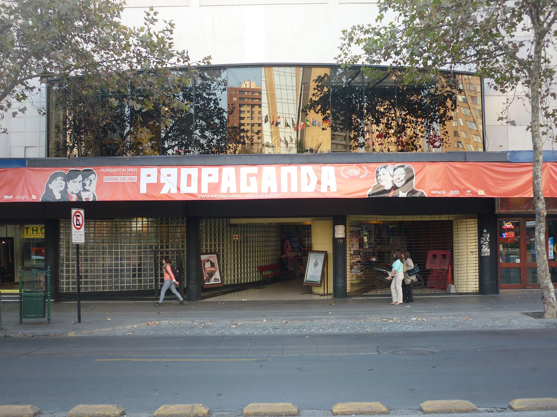 Street view of a cinema entrance with a large red Propaganda film banner, pedestrians, and trees reflected in the glass façade.