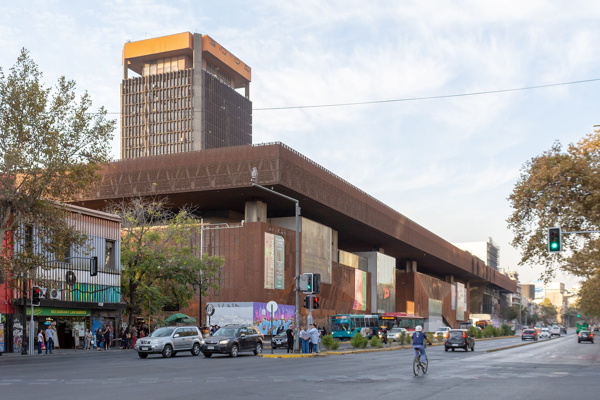 A busy city intersection with buses, cars, and a cyclist beside a long brick building and tall orange-topped tower.