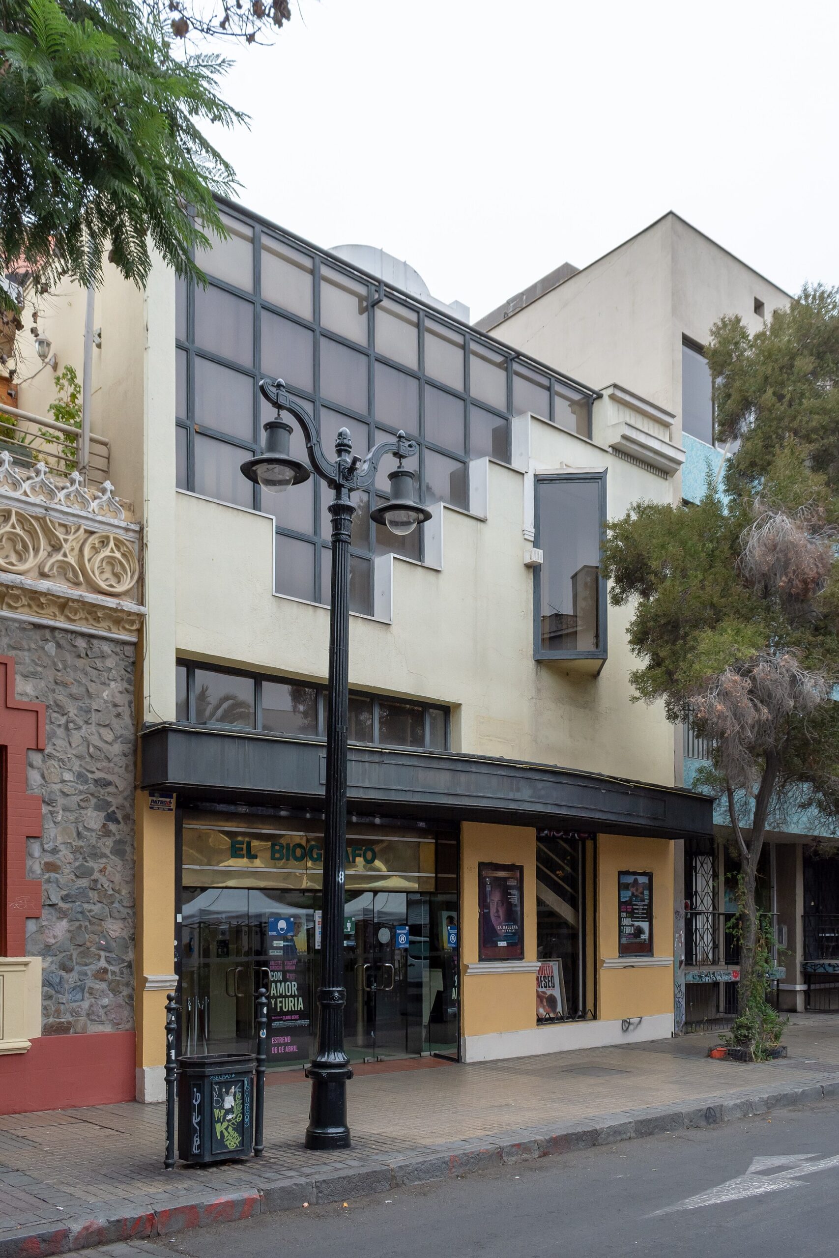 Street view of a cinema storefront beneath a modern cream-colored building, with a decorative black lamppost and trees.
