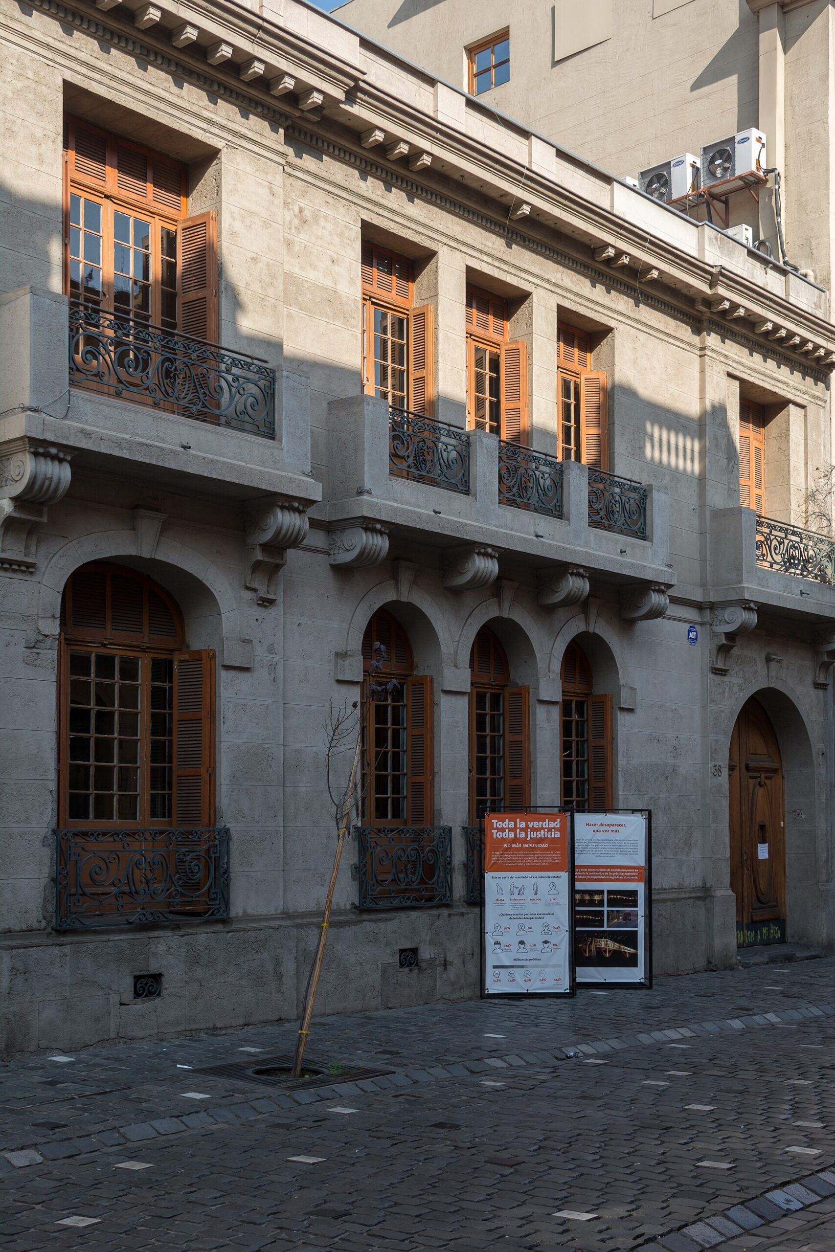 Historic stone building with arched entrances, wooden shutters, wrought-iron balconies, and public information posters on a cobblestone street.