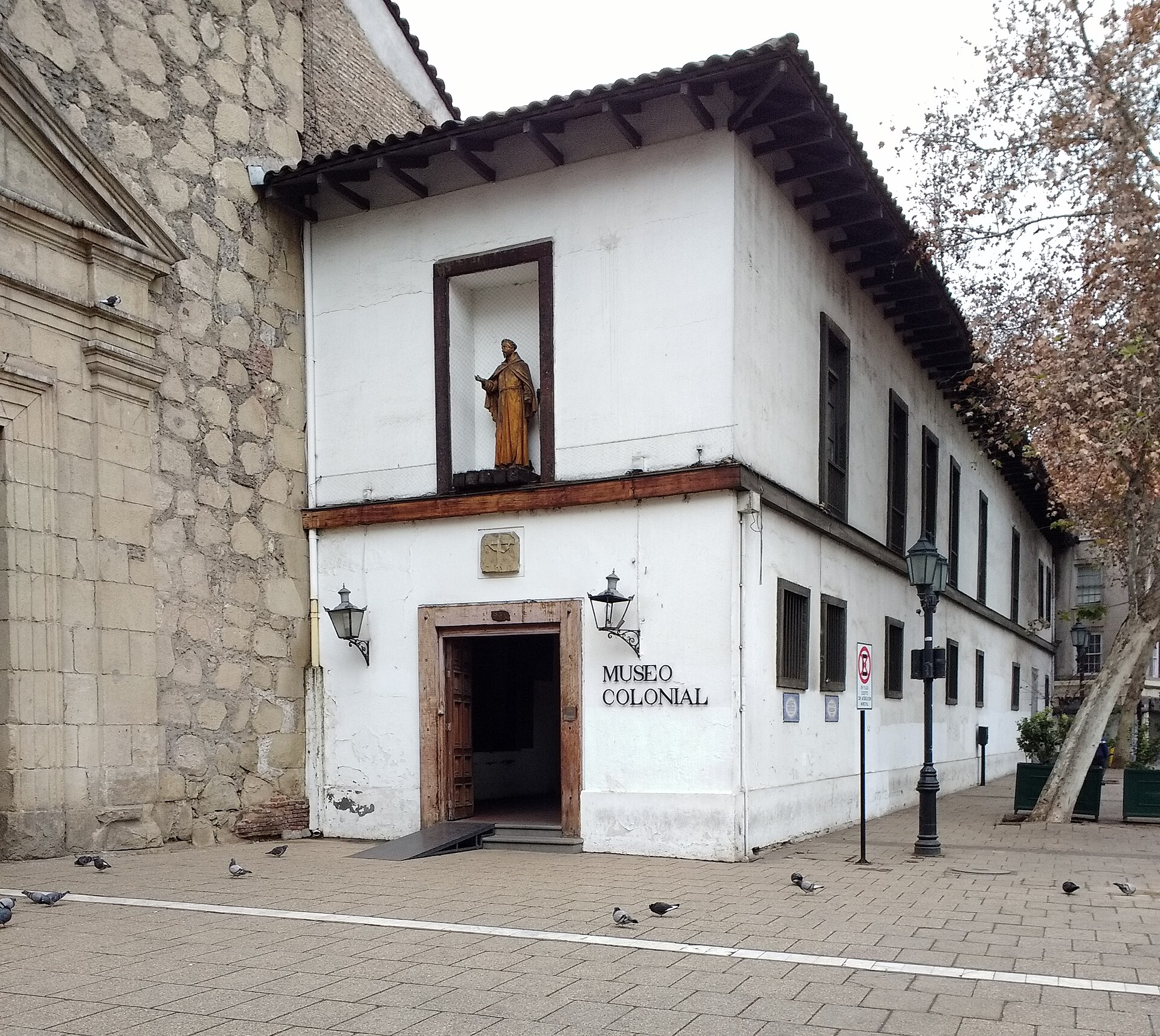White colonial-era Museo Colonial building with a statue above its entrance, beside a stone church in a pigeon-filled plaza.