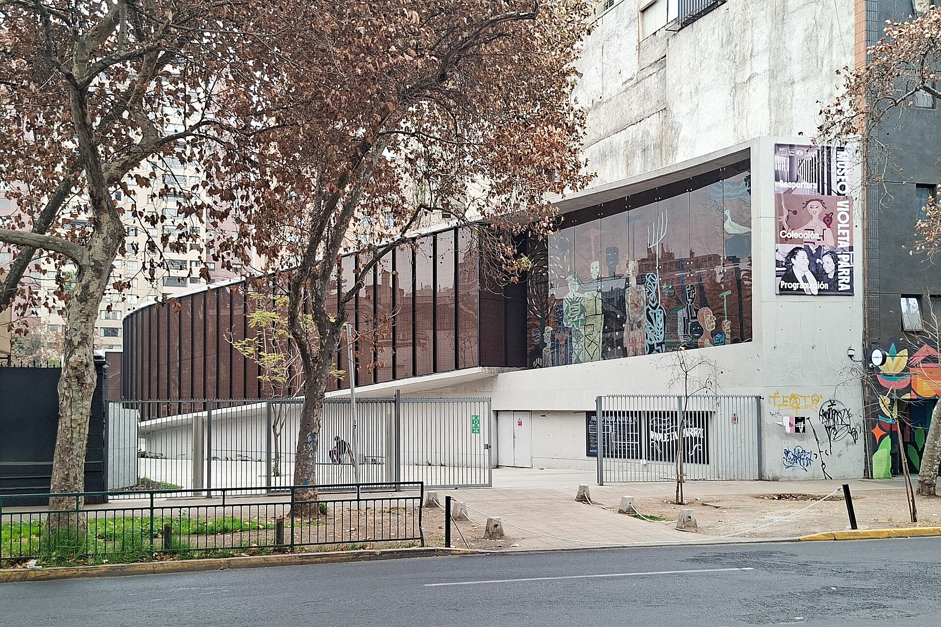 Modern museum facade with colorful artwork behind glass, Spanish-language banners, graffiti, and bare trees along the sidewalk.