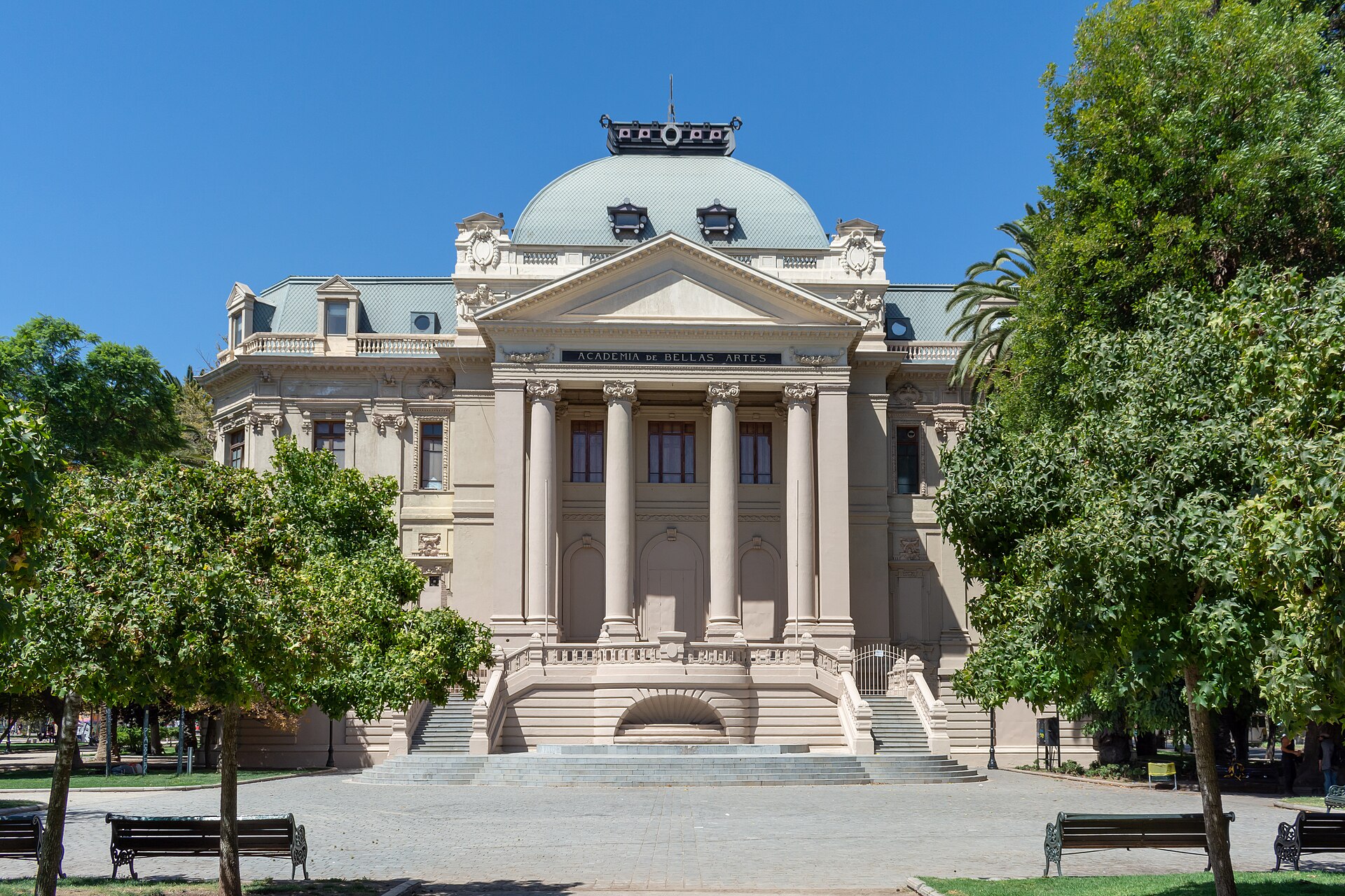 Ornate Beaux-Arts Academy of Fine Arts building with a domed roof and columned entrance, framed by trees and benches.