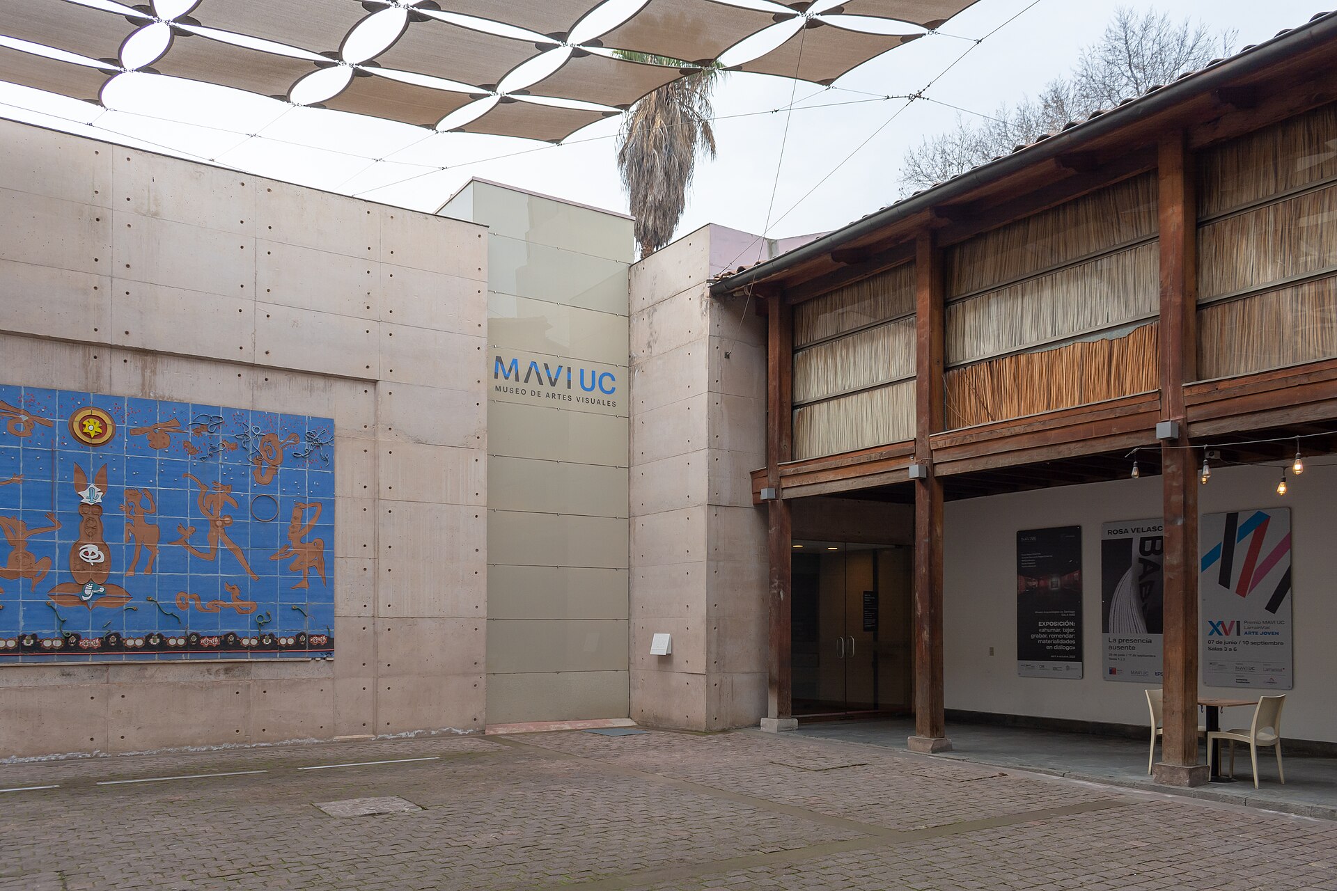 Entrance to MAVUC Visual Arts Museum, with concrete walls, a blue mural, timber facade, and overhead shade sails.