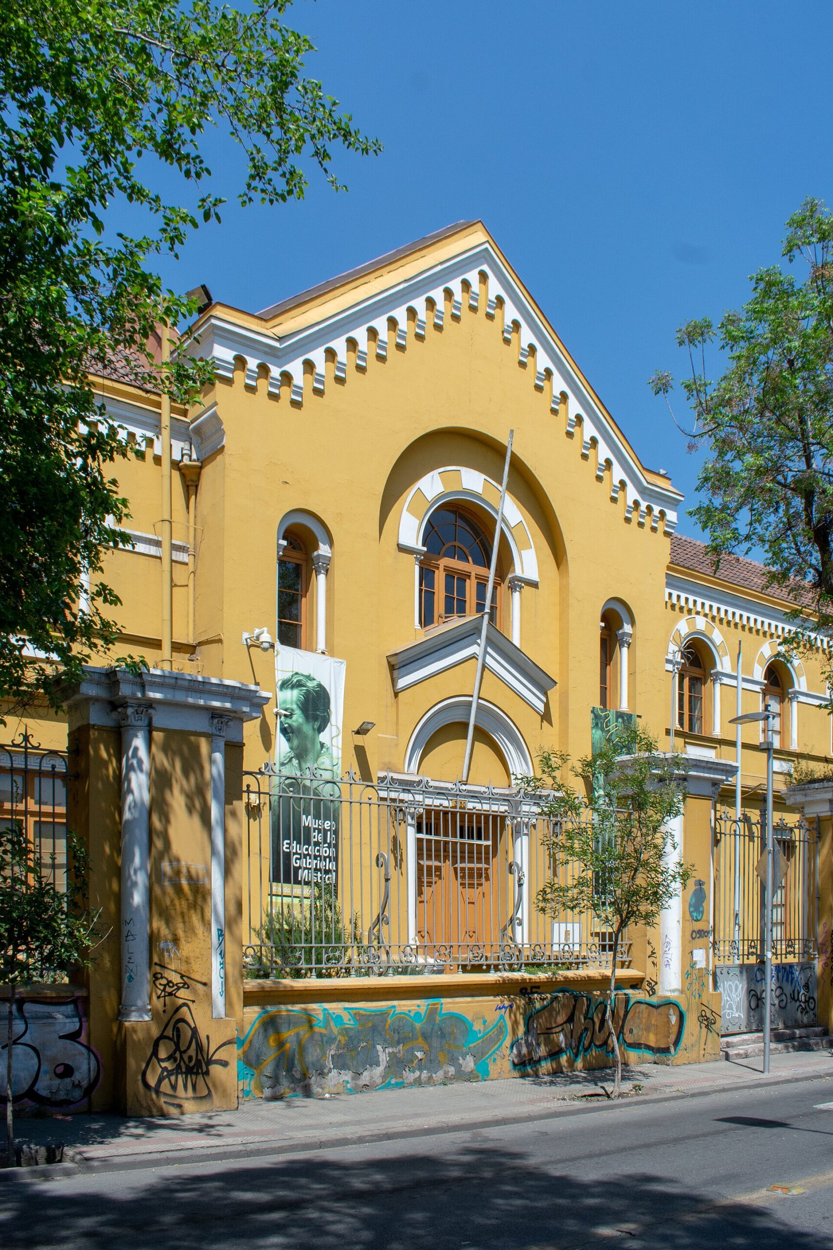 Yellow historic Gabriela Mistral education museum with arched windows, iron fence, graffiti-covered walls, and trees under a clear blue sky.