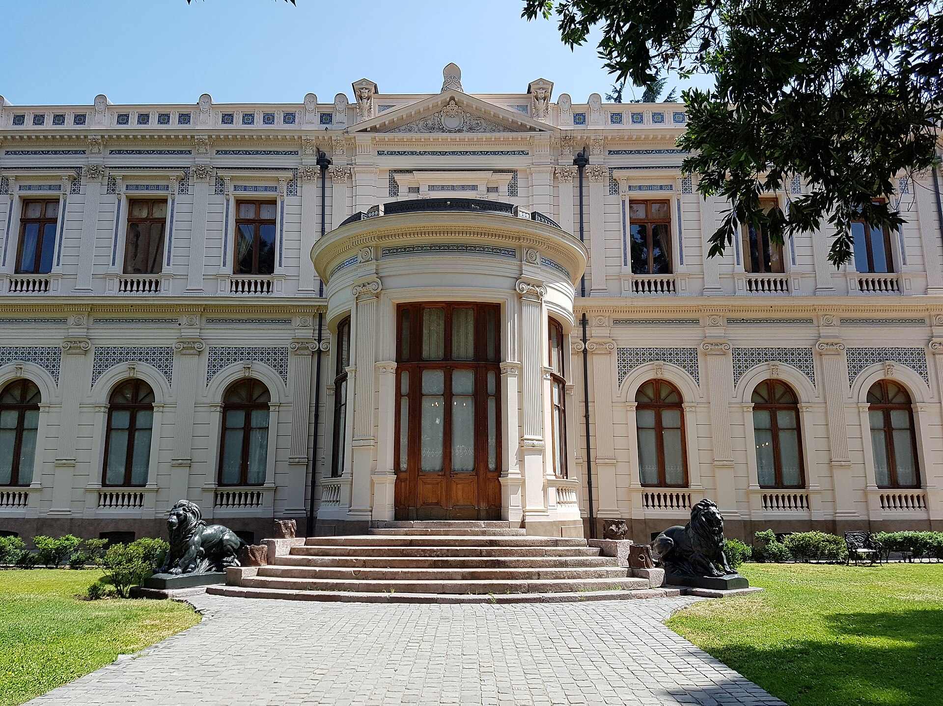 Ornate neoclassical mansion with a rounded entrance, stone steps, and lion statues in a sunny garden.